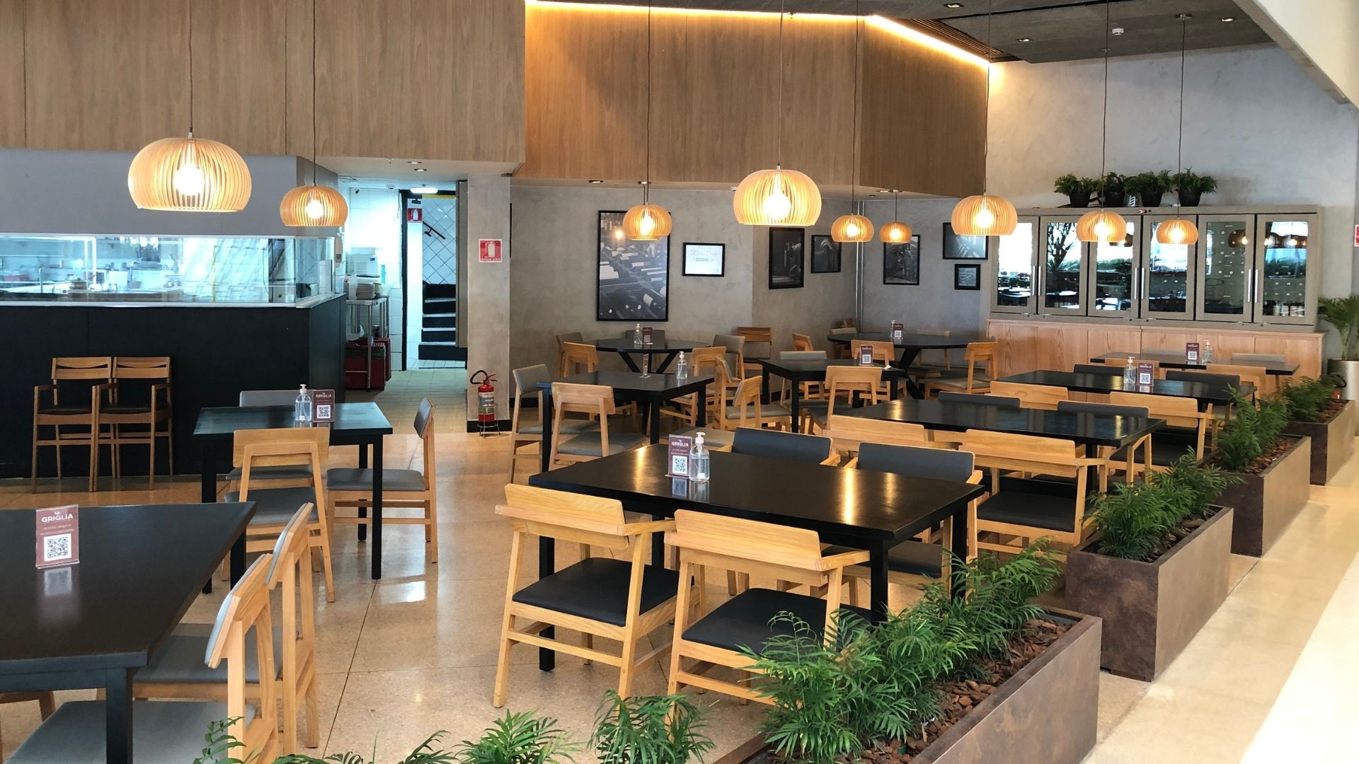Salão de restaurante moderno com mesas de madeira e tampo preto, cadeiras estofadas, luminárias pendentes em ripas de bambu, parede de concreto com quadros emoldurados, plantas verdes e vasos, iluminação quente ambiente, fotografia profissional ultra-realista