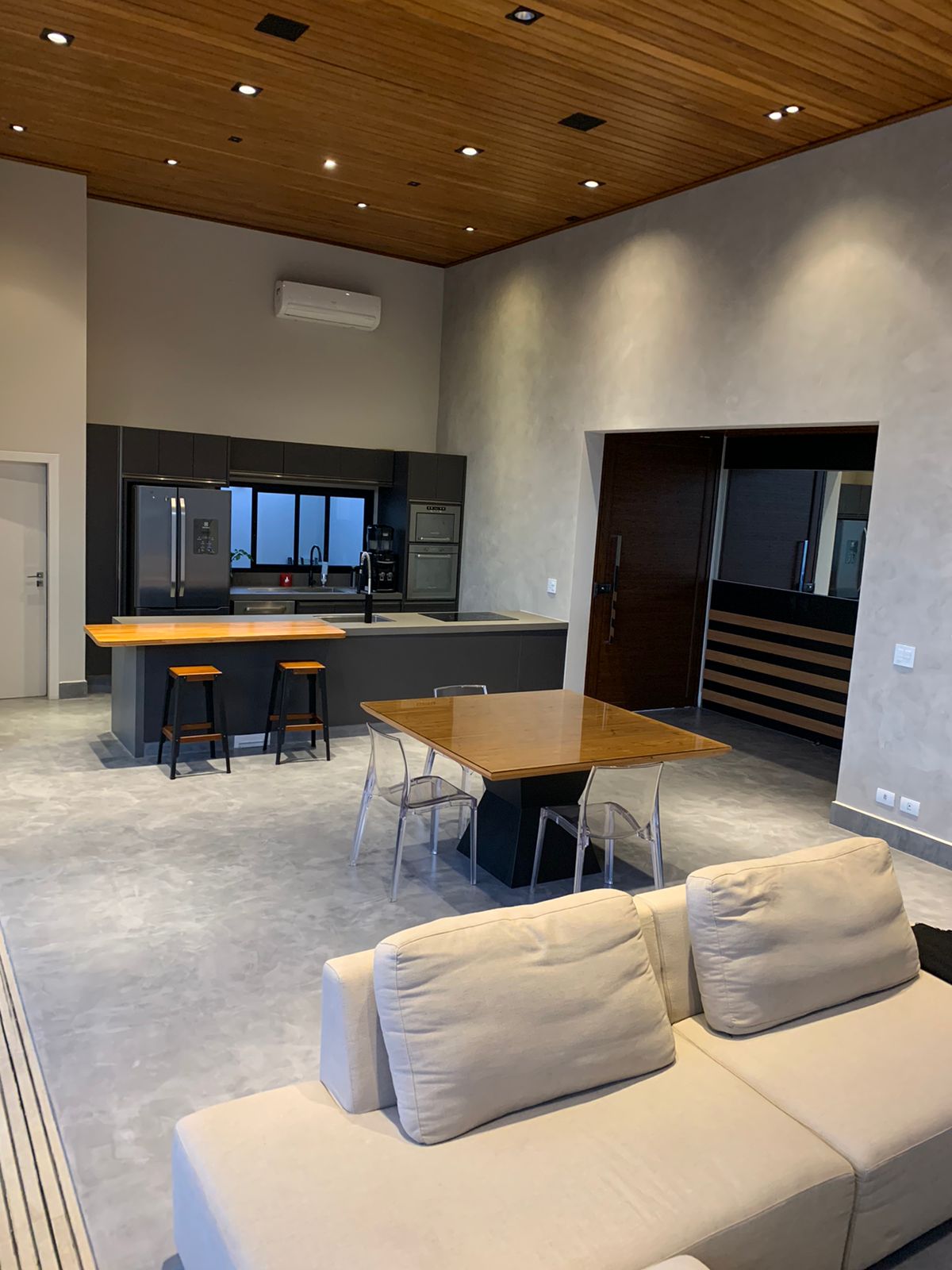 Sala de estar e cozinha integradas de reforma residencial de alto padrão, teto em ripado de madeira com iluminação embutida, marcenaria em tom grafite, piso em cimento queimado, mesa de jantar em madeira com cadeiras em acrílico transparente, sofá em linho bege, ambiente amplo e contemporâneo, fotografia realista
