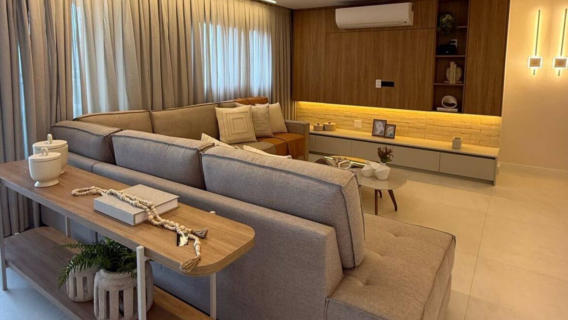 Sala de estar de apartamento reformado de alto padrão em São Paulo, sofá cinza amplo em L, almofadas texturizadas em tons neutros, marcenaria planejada em madeira com iluminação embutida, cortinas em tecido natural, decoração contemporânea aconchegante, fotografia realista com luz ambiente quente