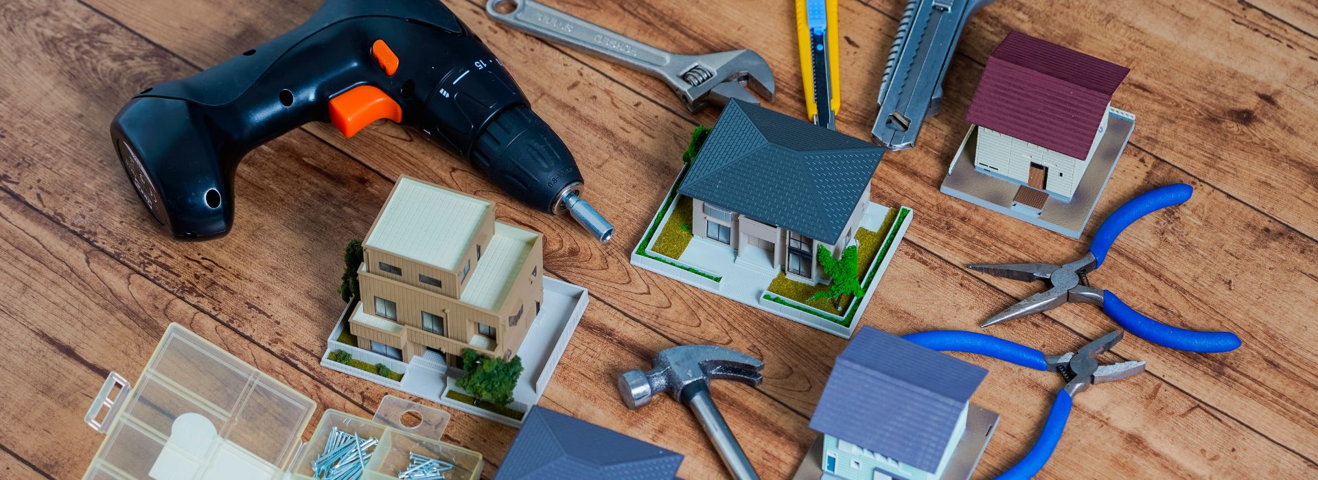 Ferramentas de construção e reforma organizadas sobre bancada de madeira, incluindo furadeira, chave inglesa, martelo e alicates, ao lado de maquetes de casas, composição plana vista de cima, tons de madeira e metal, fotografia realista