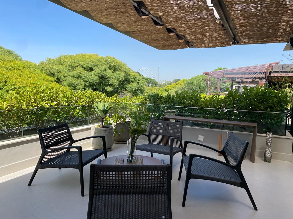 Varanda gourmet de cobertura com mobiliário em fibra sintética preta, vista panorâmica de árvores e roda-gigante ao fundo, céu azul, iluminação diurna, fotografia realista