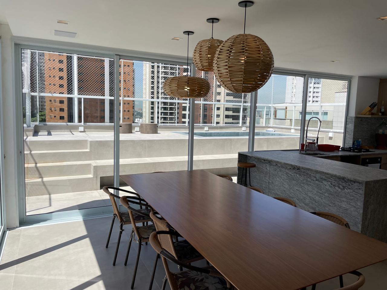 Sala de estar e jantar integrada com cozinha em apartamento de alto padrão, mesa de jantar em madeira maciça com pendentes redondos de fibra natural, janelas amplas com vista para prédios e piscina na cobertura, iluminação natural suave, fotografia de arquitetura de interiores